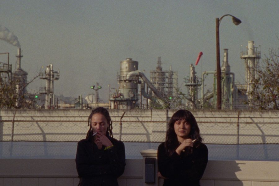 Δύο μαυροντυμένες κοπέλες καπνίζουν. Στο βάθος φαίνονται εργοστάσια.