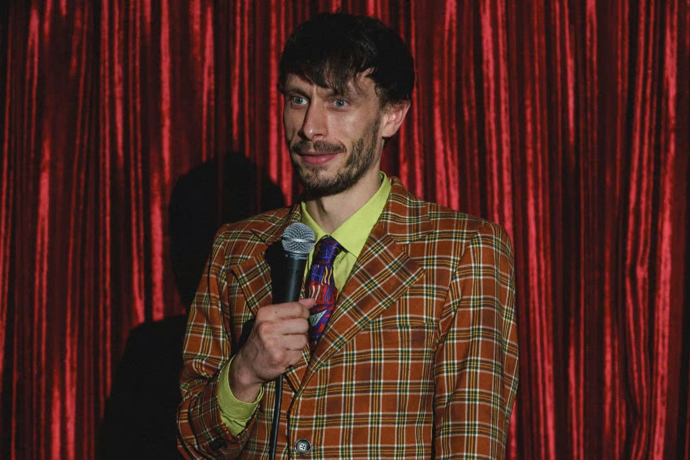 Ο Richard Gadd στο «Baby Reindeer» του Netflix. Στέκεται μπροστά από μια κουρτίνα θεάτρου, κρατώντας ένα μικρόφωνο.